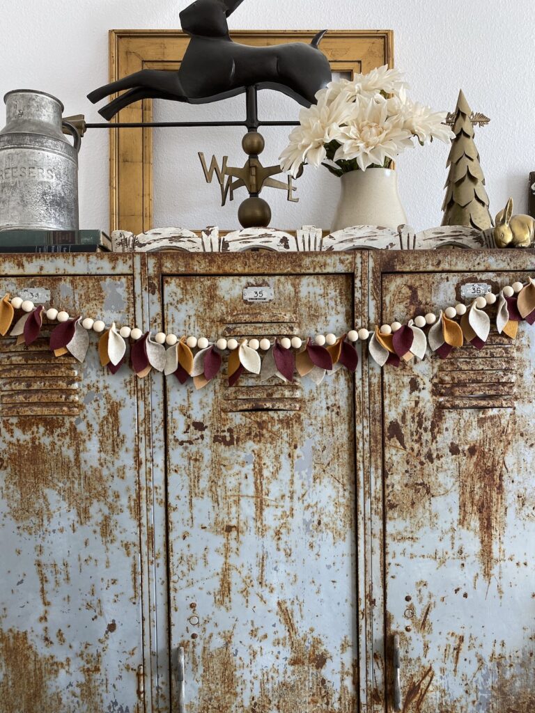 a garland of repetitive groupings of two wooden beads and a cluster of three felt leaves in fall colors such as garnet, mustard yellow, light brown, white, and grey. the garland is hung from rusted lockers being used as a buffet table. The top of the locker is decorated with more rustic farmhouse decor including a weathervane, a metal christmas tree, a vase of white dahlias, and a metal pitcher