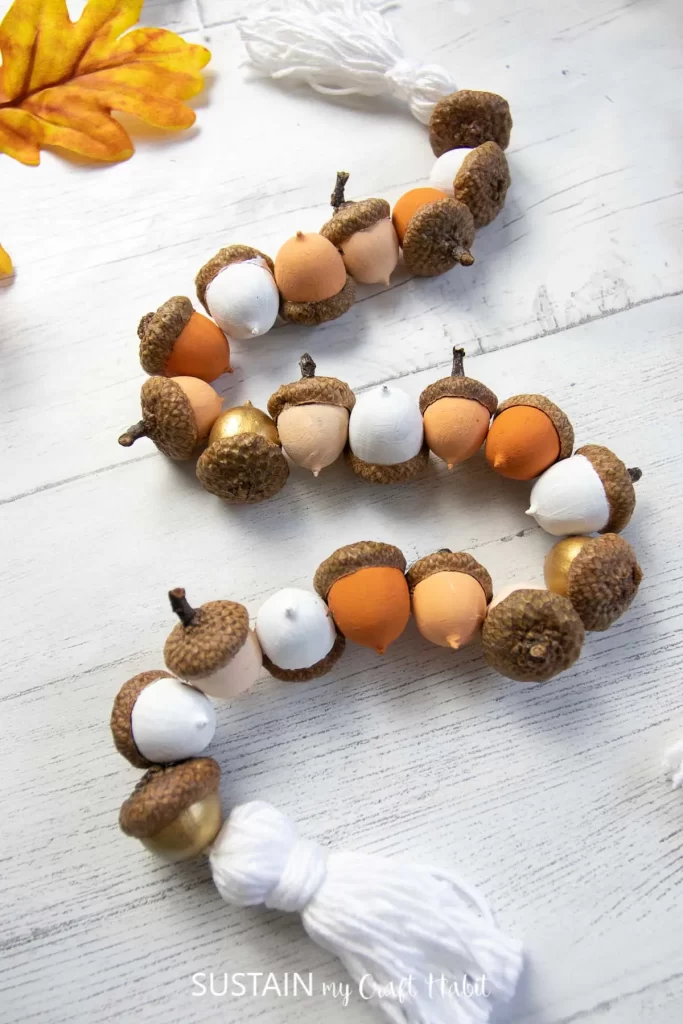 a homemade garland made with acorns strung together on a string. at both ends of the garland, there is a homemade white tassel. The acorn bodies have been painted fall colors such as orange, gold, and white white the acorn hats have been left their natural color.