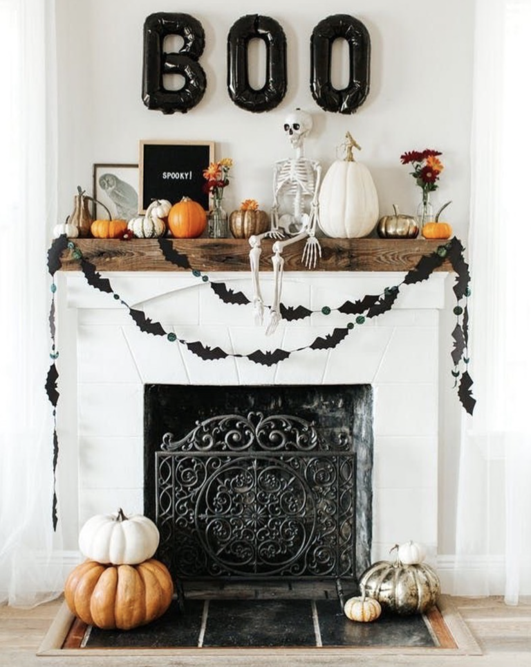 On the wall above the mantle are black letter balloons that spell "Boo". There are two garlands of black bats hanging from the mantle. Sitting on the mantle are a collection of real and fake pumpkins in colors of orange, gold, white, and green. There is a letter board that says "Spooky" and a skeleton next to some maroon flowers.