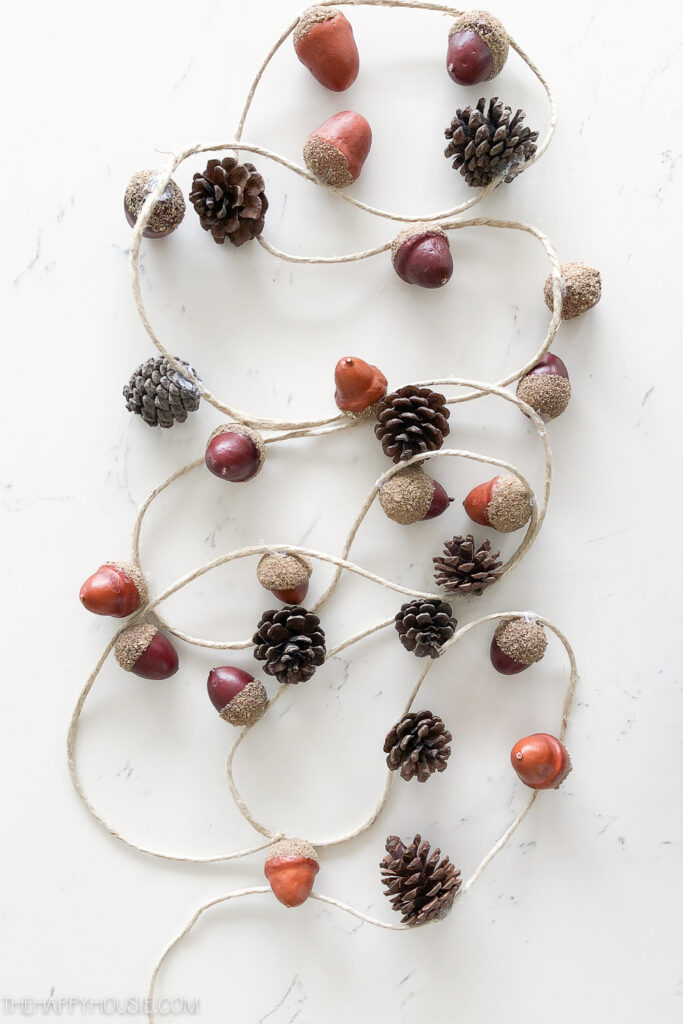 a fall garland on top of a background of a marble countertop. the garland is a jute rope with attached acorns and mini pinecones spaced in an alternating pattern. 