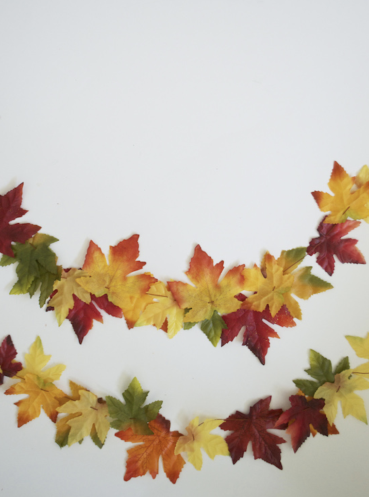 two garlands of fake fall leaves in hues of dark reds, yellows, greens, and oranges strung together with a needle and thread. the garlands are displayed against a white background. 