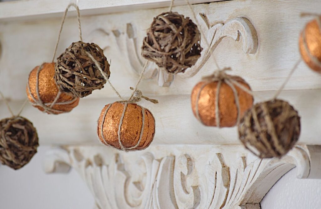 a garland of alternating mini metallic orange pumpkins and twine balls. The garland hangs from a white mantle with intricate carvings