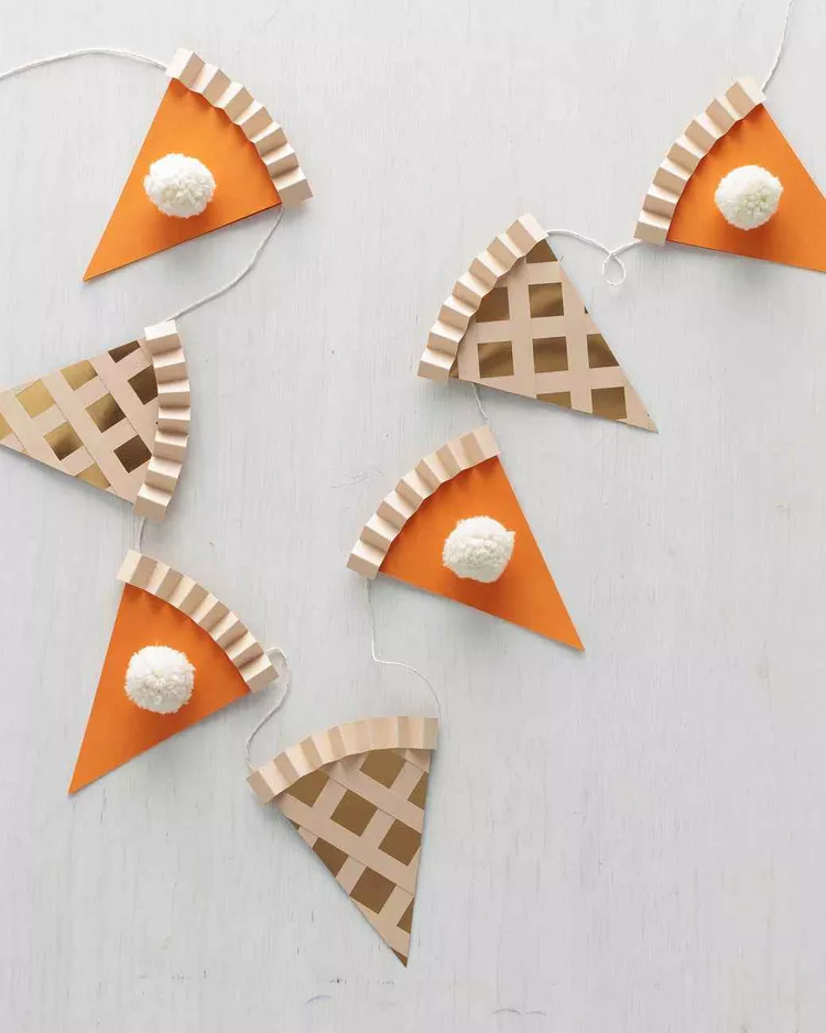 a garland of alternating pumpkin and apple pie slices made out of paper. The pumpkin pie slice uses orange card stock, has a crinkly ribbon of paper to represent the crust, and is adorned with a white pom pom to represent whipped cream. The applie pie slice is made with a gold piece of card stock. It has light brown strips on top to represent a lattice crust.