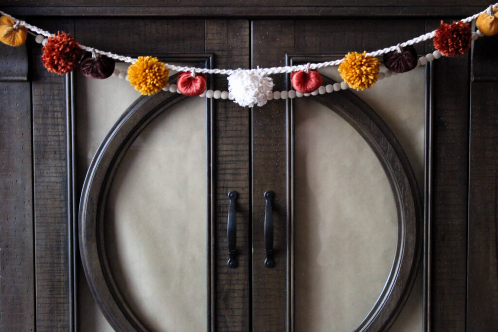 a garland of homemade yarn pom poms in alternating colors of garnet, gold, and white. they're attached to a braided white rope, which was also homemade. the pompom garland is hung over a cabinet with a strand of white beads behind it. 