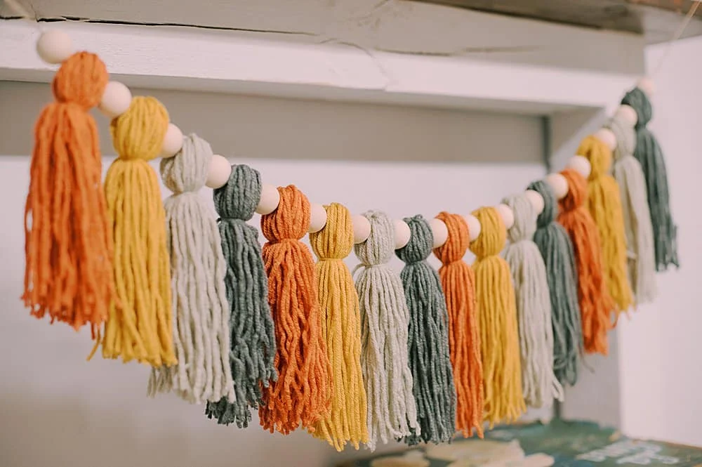 a garland of yarn tassels in colors of orange, mustard yellow, light grey, and turquoise. Each tassel is separated by a wooden bead. The garland is hung from a wooden ledge against a white background.