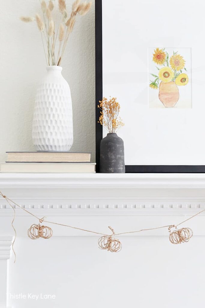 a garland of light brown wire pumpkins made from paper wrapped floral wire attached to a string of jute rope. The garland hangs from a white mantle with decor on top of it. The decor includes a white textured vase placed on top of two books and a smaller charcoal grey vase. Both vases have dried flowers in them. There's also a framed picture of a sketch of five sunflowers in a vase.